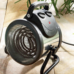 A portable electric heater with a cylindrical shape and front mesh grille sits on a tiled floor. It has a handle, two control knobs, and a red indicator light. Surrounding it are green plants.