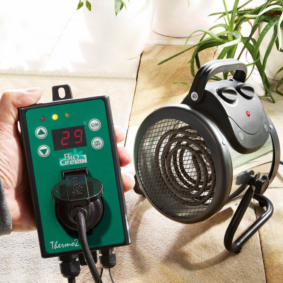 A person holds a green thermostat display.  Nearby, potted plants on tiled flooring suggest a Rhino Greenhouse setting.