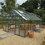 A glass Rhino Greenhouse with a green frame houses rows of potted plants on shelves, surrounded by gravel paths and trees, with a bench and large planters nearby.