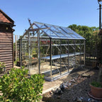 A glass Rhino Greenhouse stands on a sunlit garden patio surrounded by plants and a wooden structure with neatly arranged potted plants inside