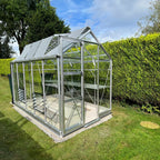 A glass Rhino Greenhouse stands with open roof vents, nestled in a neatly manicured garden, surrounded by lush green hedges and a clear blue sky above.