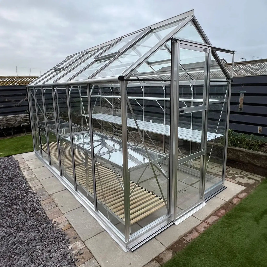 A glass Rhino Greenhouse stands idle on a paved surface, with multiple metal shelves inside. It is surrounded by a gravel path and a wooden fence in a backyard setting.