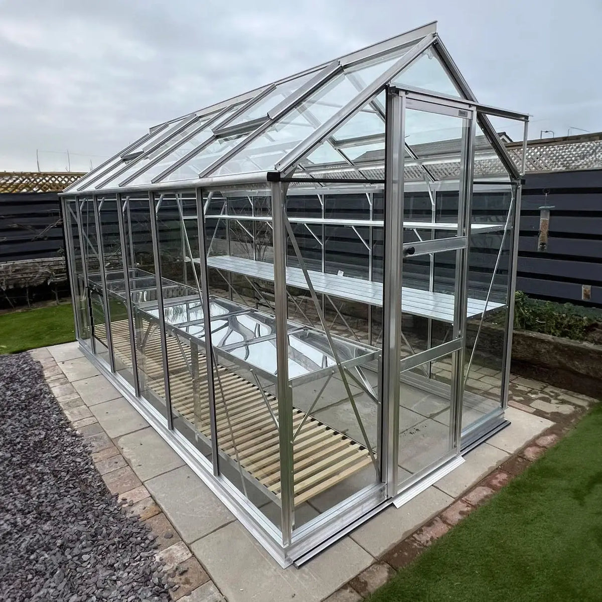 A glass Rhino Greenhouse stands idle on a paved surface, with multiple metal shelves inside. It is surrounded by a gravel path and a wooden fence in a backyard setting.