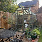 A green metal-framed Rhino Greenhouse stands in a garden, surrounded by potted plants and gravel, near patio furniture. Behind it, a brick house features rooftop solar panels.