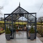 A glass Rhino Greenhouse stands open, revealing potted plants and gardening tools on shelves. It is situated outdoors, surrounded by stone walls and a rural landscape under a cloudy sky.