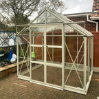 A glass Rhino Greenhouse stands on a brick patio, surrounded by a wooden fence and trees. It has a pitched roof and a visible label reading Rhino above the door.