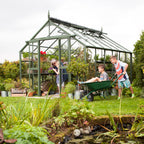 A green Rhino Greenhouse filled with plants and gardening tools stands in a lush garden. A woman tends to plants while two boys play with a wheelbarrow. Text: Rhino – Proudly British.