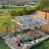 A transparent glass Rhino Greenhouse houses potted plants and chairs, situated on a stone patio. It's surrounded by a wooden fence and overlooks a lush, expansive countryside landscape.