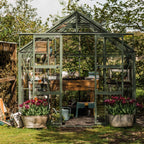 A green Rhino Greenhouse stands surrounded by lush trees, housing wooden shelves with gardening tools and pots. Two large containers of blooming tulips are positioned at the entrance.