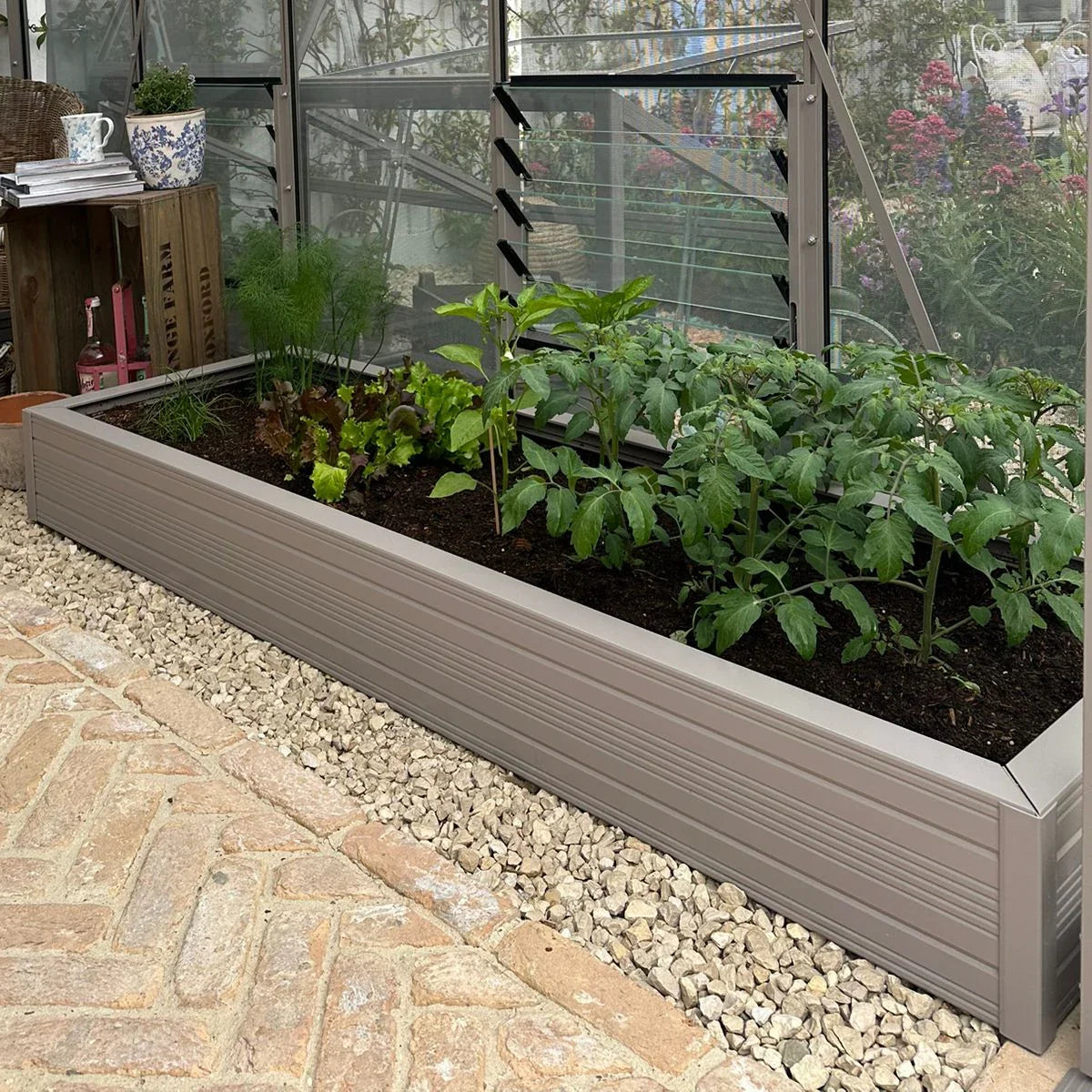 Rhino metal raised garden bed with green plants growing, set beside a Rhino Greenhouse wall amidst a gravel and brick pathway. Nearby are crates, a teapot, and garden tools.