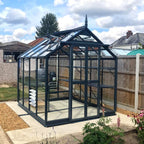 Rhino Greenhouse stands on a concrete slab, surrounded by a garden with various plants. It features glass panels and a pointed roof, enclosed by a wooden fence under a partly cloudy sky.