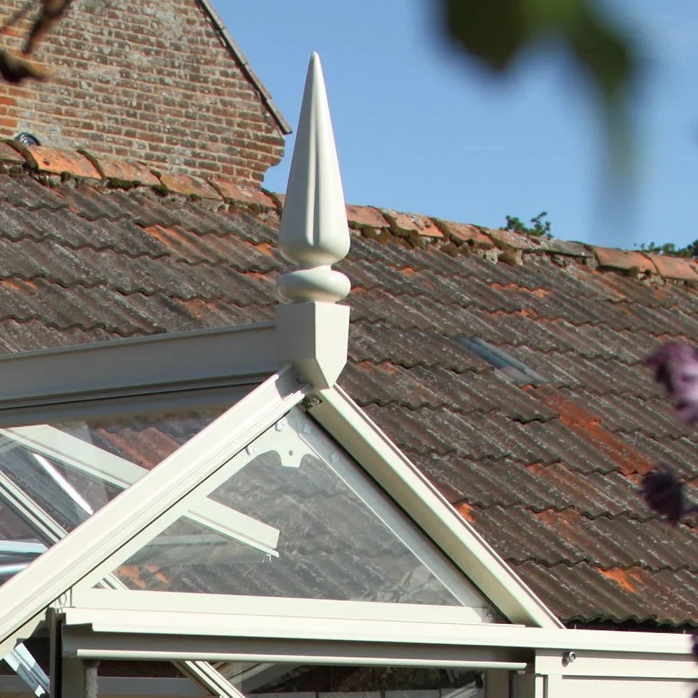 A decorative finial sits atop the glass roof of a Rhino Greenhouse, positioned against a backdrop of an old, weathered brick building with a tiled roof under a clear blue sky.