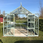 Rhino Greenhouse with green metal frame and glass panels stands on grass. Inside, shelves and tables are arranged. Surrounding are trees and a blue sky, creating a serene garden atmosphere.
