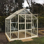 A glass and metal Rhino Greenhouse stands neatly on a wooden base, framed by trees and bushes, with visible interior plant beds and a sliding door for entry.