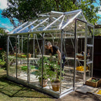 A glass Rhino Greenhouse shelters a person tending to plants in pots. Inside, gardening tools and containers are visible on shelves. It's set outdoors in a sunny garden with trees.