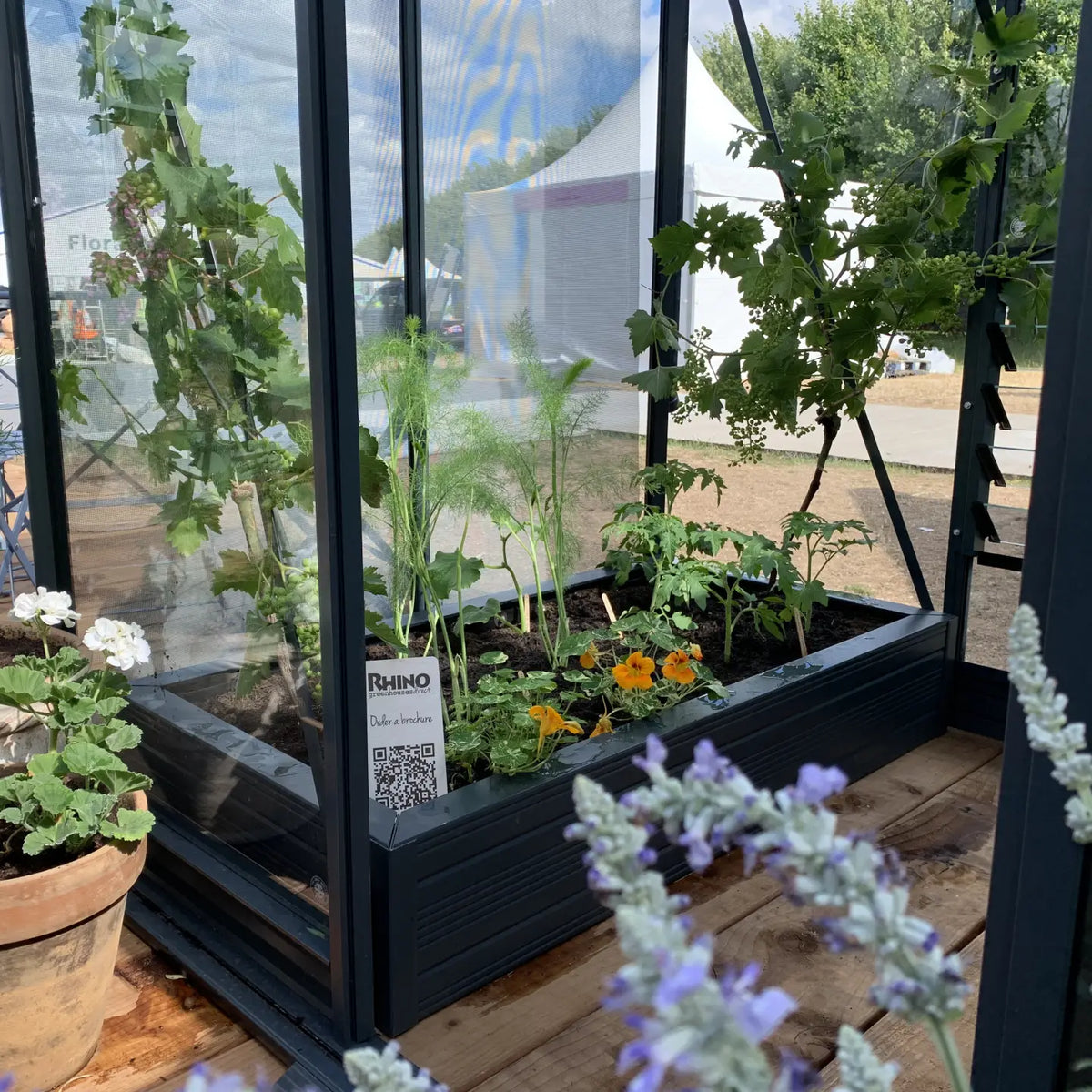 A Rhino Greenhouse contains various plants growing in planter boxes, surrounded by a vibrant garden setting. 
