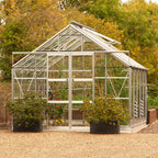 A glass Rhino Greenhouse filled with plants, including climbing tomatoes, stands on a gravel path. It is surrounded by autumn foliage and features two planter boxes nearby.