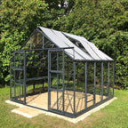 A glass and metal Rhino Greenhouse stands empty on a concrete slab, surrounded by lush green foliage. Sunlight filters through its transparent panels, highlighting its sturdy, modern structure.