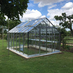A glass-paneled Rhino Greenhouse stands on a grassy lawn, surrounded by trees and shrubs, under a blue sky with fluffy clouds.