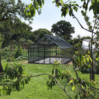 A Rhino Greenhouse stands on a lawn, surrounded by trees and garden plants under a clear sky. It's centrally positioned, with visible structural details and an adjacent raised bed.