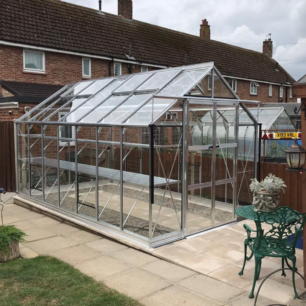 A glass Rhino Greenhouse with a metal frame stands on a patio near residential brick houses, surrounded by a wooden fence. Nearby are green metal garden furniture and potted plants.