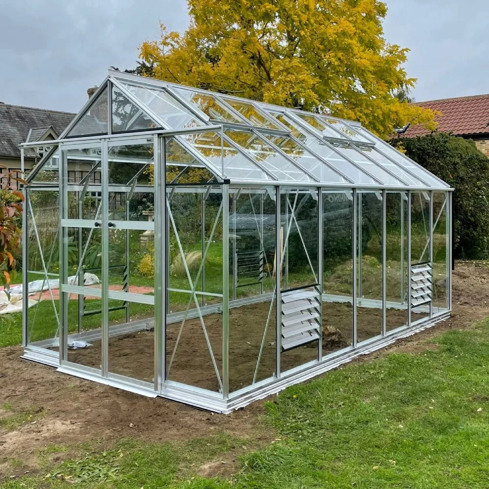 A glass Rhino Greenhouse with a metal frame stands on a patch of bare soil, surrounded by grass and autumn trees with yellow leaves in a residential area.