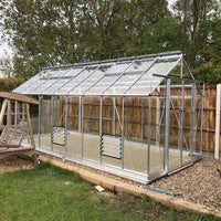 A glass Rhino Greenhouse stands on a gravel foundation, with a wooden fence and trees in the background. It features metal framing and adjustable vents, situated on a neatly maintained lawn.