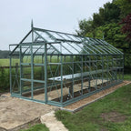 A large glass Rhino Greenhouse stands on a gravel foundation with a metal frame, set amidst a rural garden with lush greenery and trees in the background.