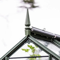 A green Rhino Greenhouse roof with decorative finial shows leaves inside, framed by wet glass panes. A tube structure is attached, against a blurred white background.