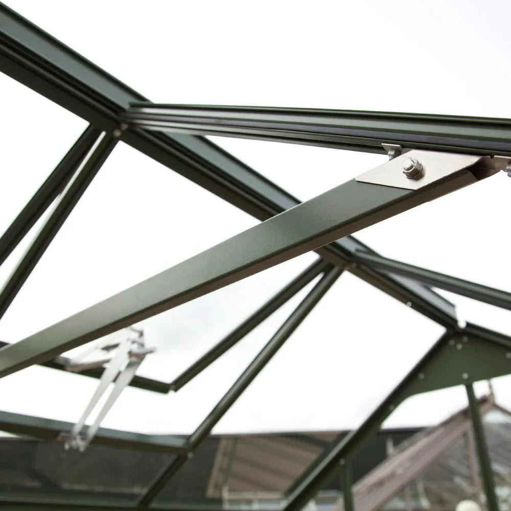 A dark green metal Rhino Greenhouse frame is shown from below, with connected beams and bolts, set against an overcast sky.