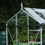 The Rhino Greenhouse has a pointed roof and transparent panels, housing potted plants. It's surrounded by dense green foliage, with sunlight illuminating the structure's metal framework.