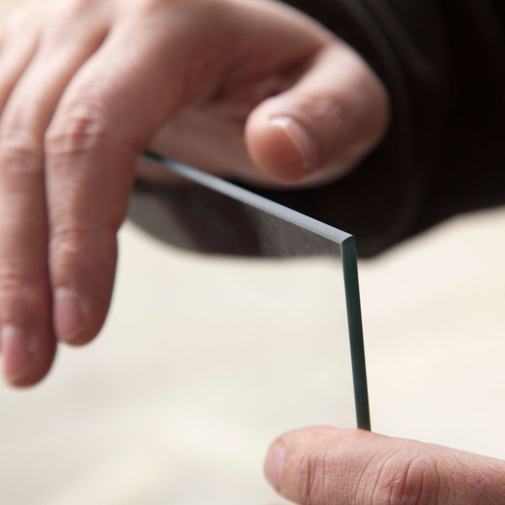 Hands hold a thin, rectangular piece of glass vertically, displaying its clarity and smooth edges against a soft, blurred background.