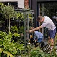 A metal-framed Rhino Greenhouse showcases plants while a man and child interact with it. They are in a lush garden, surrounded by various greenery and near a building entrance.
