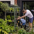 A metal-framed Rhino Greenhouse showcases plants while a man and child interact with it. They are in a lush garden, surrounded by various greenery and near a building entrance.