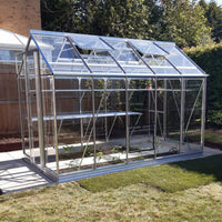 A glass Rhino Greenhouse with open windows sits on a paved area, surrounded by grass and trees, with some plants growing inside.
