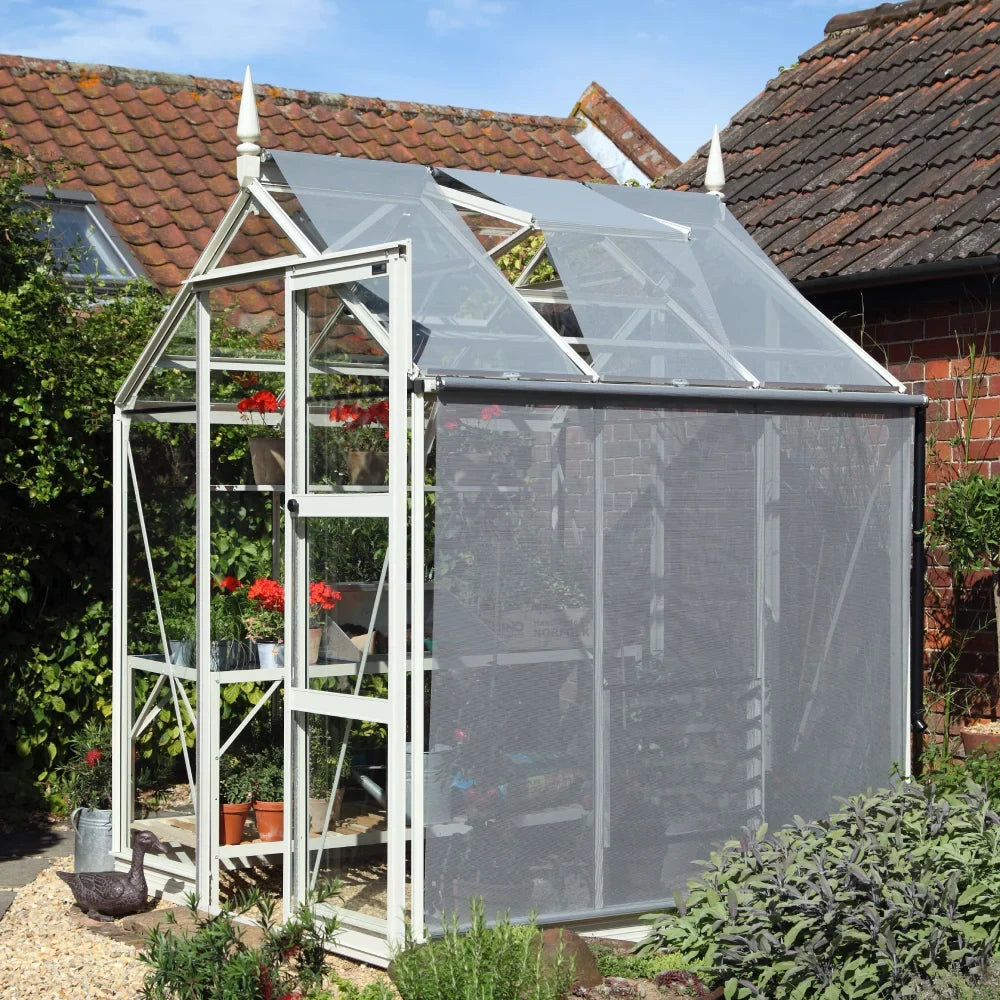 A white-framed glass Rhino Greenhouse stands outdoors, filled with potted plants and flowers. It is adjacent to brick and tile-roofed structures, surrounded by lush garden greenery under a clear sky.