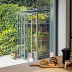 Rhino Greenhouse displaying vibrant plants, located in a garden with lush greenery. Viewed through a window, a wooden table holds pastries on a board next to mugs and a coffee pot.